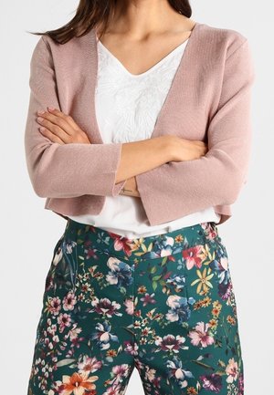 Cardigan rosa chiaro con design corto e maniche a tre quarti, indossato sopra una maglietta bianca ricamata e pantaloni a stampa floreale.