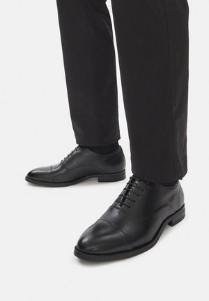 Schwarze Lederschuhe mit glatter Oberfläche, abgerundeter Spitze und Schnürdesign, kombiniert mit schwarzen Hosen.