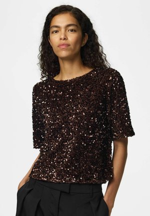 Femme aux cheveux bouclés portant un haut à manches courtes marron foncé recouvert de sequins brillants et un pantalon noir plissé, les mains dans les poches.