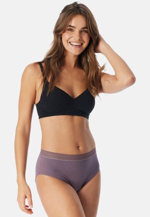 Smilende kvinde med langt brunt hår iført en sort bh og lilla høj-taljede undertøj, poserede med den ene hånd, der rørte ved hendes hår.