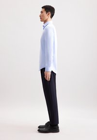 Camicia azzurra a maniche lunghe con colletto a punta, abbinata a pantaloni scuri e scarpe nere, mostrata in profilo laterale su uno sfondo semplice.