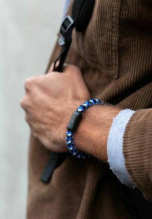 Kék és fehér fonott karkötő matt fekete záródással. Karon viselve, kordbársony kabáttal és látható ingmelllel párosítva.