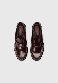 Bourgondische leren instappers met een gladde afwerking, voorzien van een gestikte detail rond de zool en een kleine metallic accent aan de voorkant.