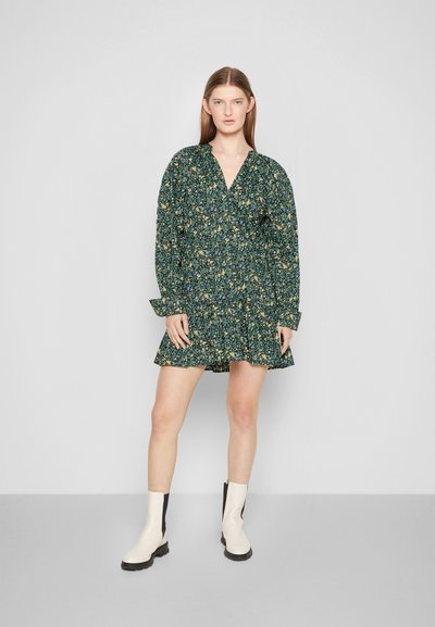 Vestido floral verde escuro com mangas longas e decote em V. Apresenta uma saia rodada e um corte solto. Combinado com botas de cano curto em creme.