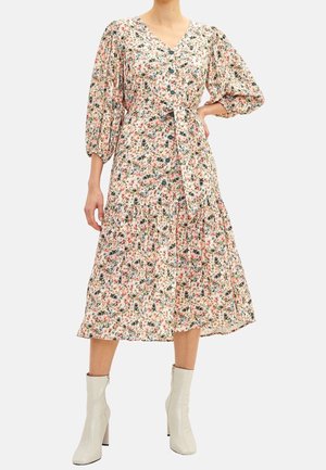 Femme portant une longue robe fleurie avec des boutons à l'avant, des manches bouffantes et une taille nouée, associée à des bottines claires.