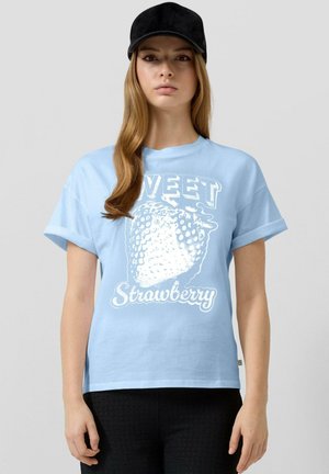 Jeune femme portant un t-shirt bleu clair avec un grand motif de fraise blanche et le texte « Sweet Strawberry », ainsi qu'une casquette noire.