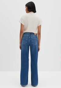 Wijduitlopende blauwe jeans met een hoge taille, achterzakken en een bruine merkpatch. Gecombineerd met een lichtbeige ribbelachtig T-shirt met korte mouwen.