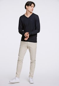 Maglione a V blu navy in tessuto morbido, abbinato a pantaloni slim-fit beige chiaro e sneakers bianche. Design semplice, senza motivi visibili.
