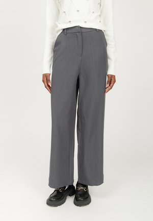 Persoon in een grijze wijde broek, witte trui met parel details en zwarte leren loafers met gouden kettingen, staand op een witte achtergrond.