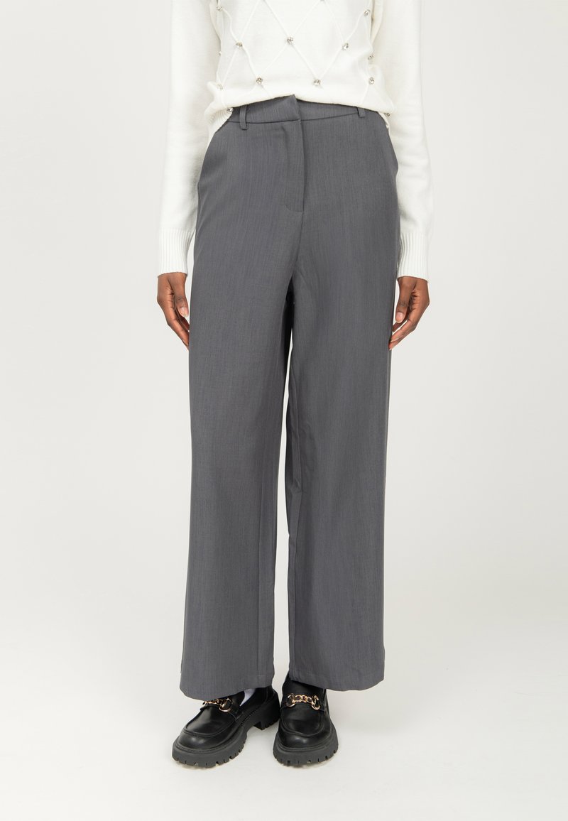 Persona vestida con pantalones anchos grises, suéter blanco con detalles de perlas y mocasines de cuero negro con cadenas doradas, de pie sobre un fondo blanco.