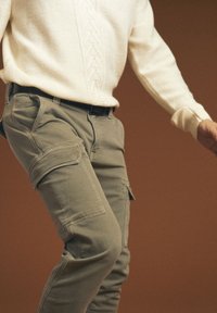 Osoba ubrana w beżowe spodnie cargo i kremowy sweter z dzianiny na brązowym tle, w ruchu.