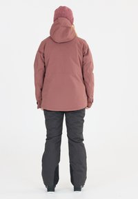 Wasserdichte weinrote Jacke mit Kapuze, kombiniert mit schwarzen isolierten Hosen und dunklem Schuhwerk, von hinten auf einem weißen Hintergrund gesehen.