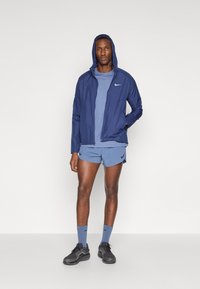 Men's athletic wear: navy hooded jacket, light blue textured short-sleeve top, matching shorts. Accessories include blue socks and dark sneakers.