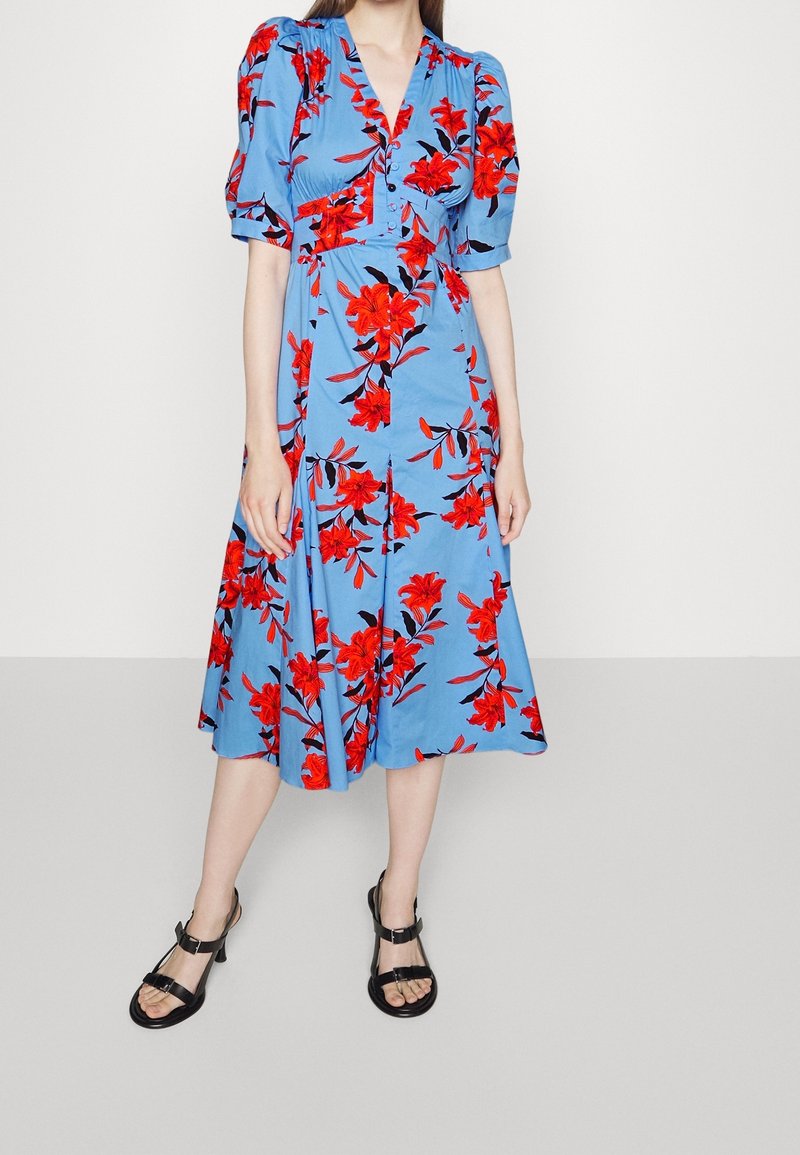 Robe fleurie bleue avec des motifs de fleurs rouges et noires, manches courtes bouffantes, détails à boutons et jupe évasée. Portée avec des sandales noires.