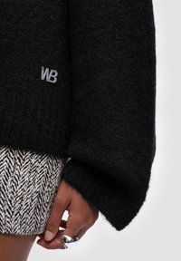 Czarny wełniany sweter o teksturowanym wykończeniu, z małym szarym logo "WB". W zestawie ze spódnicą w wzór jodełki, prezentującą kontrastujące wzory.