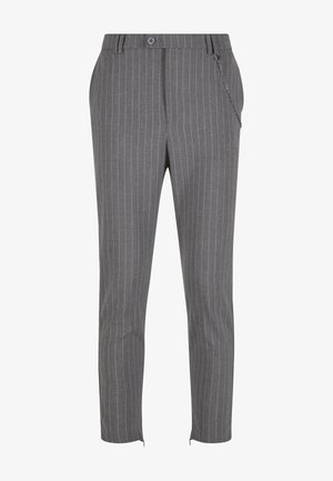 Férfi szürke tűcsíkos, testreszabott nadrágban lánc kiegészítővel és fehér tornacipővel, sík háttér előtt áll.