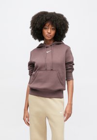 Barna Nike kapucnis pulóver elülső zsebbel, húzózsinórokkal és fehér logóval. Bézs melegítőnadrággal párosítva, laza sportos stílust mutatva.