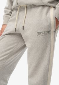 Gray sweatshirt pants made of soft fabric. Features a light cream side stripe and a printed logo in dark gray on the left thigh.