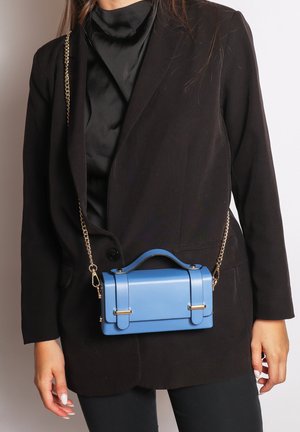 Vrouw in zwarte blazer en coltrui met een kleine blauwe leren handtas met een gouden kettingband over het lichaam.