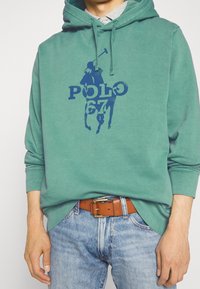 Grön hoodie med en blå "POLO 67" grafik. Tillverkad i mjukt material, den har en känguruficka och ribbade muddar, kombinerad med ljusblå jeans.
