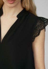 Černá halenka s hlubokým výstřihem do V, zdobená krajkovými detaily na ramenou. Měkká tkanina s hladkou strukturou dodává designu jemný nádech.
