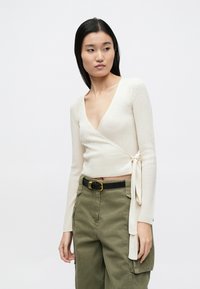 Top crem, cu model nervurat și închidere cu cordon, mâneci lungi, asortat cu pantaloni verde măsliniu cu talie înaltă și curea neagră.