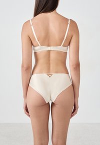 Femme vue de dos portant un soutien-gorge en satin beige et une culotte taille haute assortie avec un petit détail en découpes décoratives.