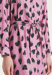 Vestido rosa con patrones de fresas en negro y verde, que presenta un frente abotonado, mangas largas y un cinturón atado en la cintura.