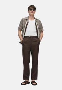 Short-sleeve patterned shirt in brown and cream, white tank top, brown linen pants, and brown flip-flops, with dark sunglasses.