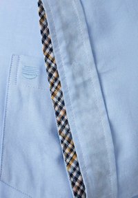 Jasnoniebieska koszula o jednolitej fakturze, z kratkowanym akcentem wewnątrz kołnierza i w okolicy kieszeni.