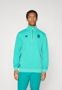 Φούτερ σε φωτεινή τιρκουάζ απόχρωση, με σχεδιασμό μισής φερμουάρ, κεντημένα λογότυπα και ριγωτές μανσέτες και τελείωμα. Μαλακή υφή υφάσματος.