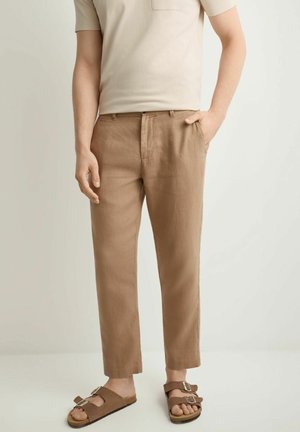 Mężczyzna ubrany w beżową koszulę z krótkim rękawem, jasnobrązowe spodnie i brązowe sandały z klamrami, stojący z jedną ręką w kieszeni.