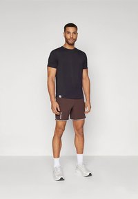 Camiseta escura masculina e calções castanhos com borda branca e logótipo. Os calções têm comprimento médio e a camisola apresenta um decote redondo clássico.