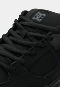 Crne tenisice za skejtanje s teksturiranim sintetičkim gornjim dijelom, ravnim vezicama, podstavljenim jezikom i suptilnim plavim naglaskom uz vanjski rub potplata. Logotip DC na jeziku.