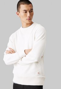 Felpa leggera color crema realizzata in tessuto morbido, con scollatura rotonda e polsini a coste. Dettaglio del piccolo logo sull'orlo e una vestibilità ampia.