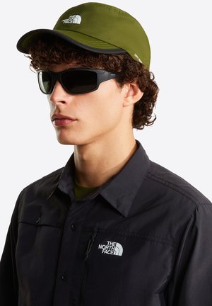 Grüne Mütze mit schwarzem Schirm und weißem Logo, schwarze Sonnenbrille und dunkles Hemd mit weißem Logo auf der Brust, strukturiertes Gewebe.