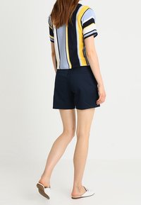 Kortærmet, stribet skjorte i blå, gul og marineblå, parret med mørke shorts. Modellen har hvide slip-on sko på.