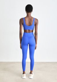 Femme se tenant de dos, portant un soutien-gorge de sport bleu, un legging taille haute assorti et des baskets blanches, sur un fond uni.