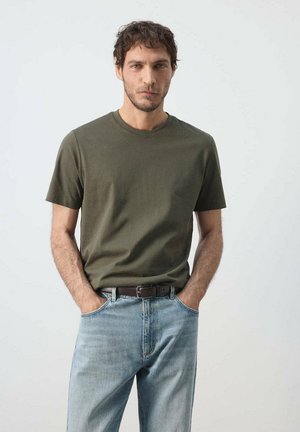 Mann mit dunklen Haaren in olivgrünem T-Shirt und hellblauen Jeans steht mit den Händen in den Taschen vor einem einfarbigen Hintergrund.
