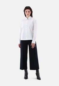 Camicia bianca con colletto e bottoni frontali, abbinata a pantaloni larghi blu scuro e stivali neri alla caviglia. Tessuto morbido, vestibilità sartoriale.