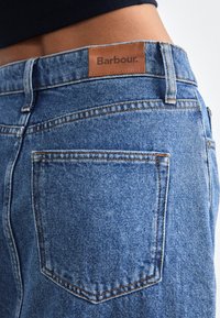 Sinised denim teksad, millel on nahkplaaster, millel on kiri "Barbour." Omab kahekordset õmblust ja tagakotti, millel on nurgeline disain.