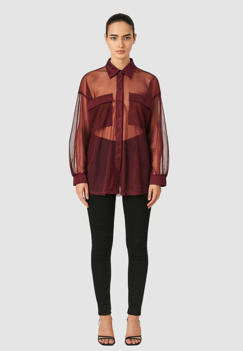 Camicia trasparente bordeaux con bottoni, maniche a sbuffo lunghe e due tasche anteriori, indossata con pantaloni neri aderenti e sandali con apertura sulla punta.