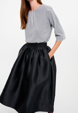 Femme portant un haut argenté en sequins à manches trois-quarts, rentré dans une jupe midi noire en satin taille haute, les mains dans les poches.