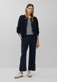 Cardigan a coste blu navy con scollatura rotonda e chiusura a bottone, abbinato a una camicia a righe a maniche lunghe e pantaloni larghi blu navy. Sneakers bianche.