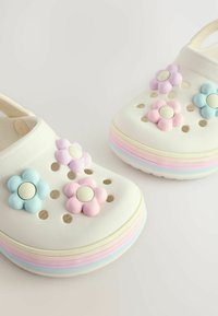 Zoccoli bianchi con suole arcobaleno pastello, decorati con charm a forma di fiore nei colori rosa, viola e blu sulla parte superiore.