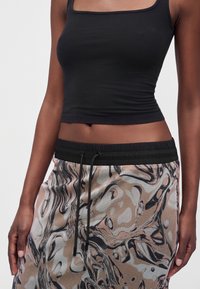 Femme portant un crop top noir et un pantalon à cordon avec un motif abstrait tourbillonnant beige, noir et blanc, debout devant un fond uni.