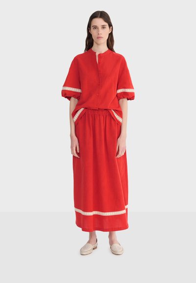 Femme debout portant un chemisier rouge boutonné avec des manches bouffantes et une jupe longue assortie, toutes deux ornées de dentelle blanche, et des chaussures beiges sans lacets.