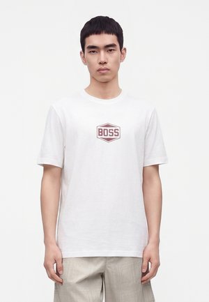 Junger Mann mit kurzen schwarzen Haaren, der ein weißes T-Shirt mit rotem "BOSS"-Logo und hellbeige Hose trägt, steht vor einem einfarbigen Hintergrund.