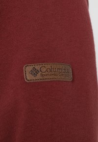 Sweat-shirt bordeaux à texture lisse, avec un patch en cuir marron embossé du texte "Columbia Sportswear Company."