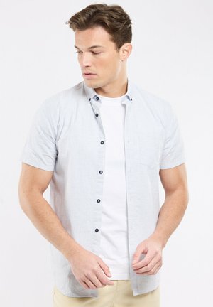Homme portant une chemise à manches courtes bleu clair par-dessus un t-shirt blanc, pantalon beige, regardant vers le bas et à gauche sur un fond uni.